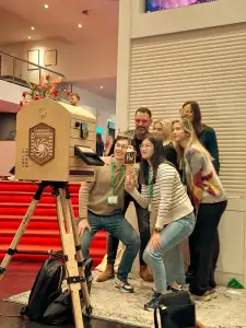 Zeven mensen poseren op een evenement in het Beatrix Theater in Utrecht, voor een vintage fotohokje met een rode loper als achtergrond. Eén persoon houdt een bordje met 'FW' vast. De setting is binnen met moderne decoratie.