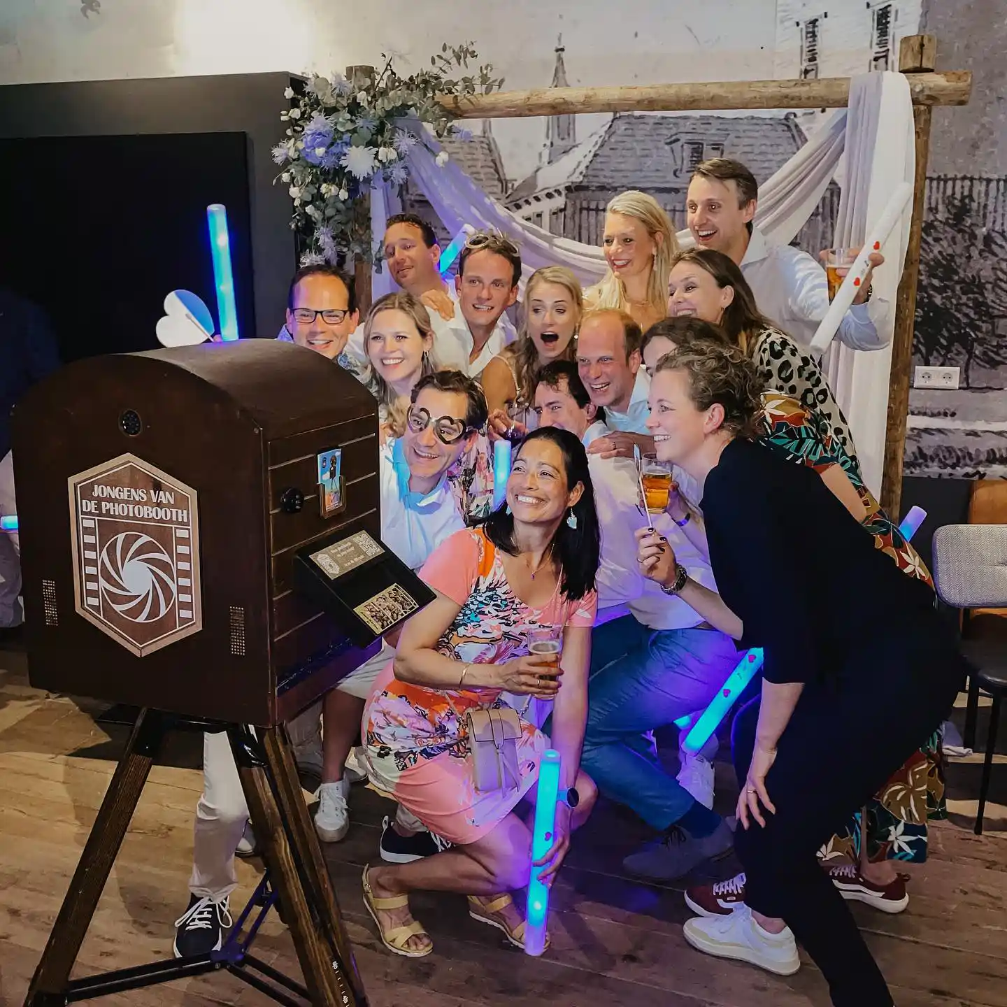 gastenboek photobooth foto van een groep feestgasten die lachend poseren bij een vintage fotohokje, omringd door lichtgevende sticks en een vrolijke sfeer.